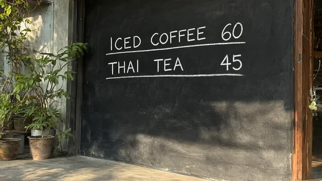 Qwen-Image 2.1: A hand-written chalkboard menu outside a small Bangkok cafe that reads "ICED COFFEE 60" and "THAI TEA 45" in neat white chalk letters, potted plants and morning sun, photograph
