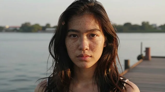 Z-Image Turbo: A close portrait of a young Thai woman with freckles and loose dark hair on a riverside pier at golden hour, warm rim light, shallow depth of field, natural skin texture, photograph
